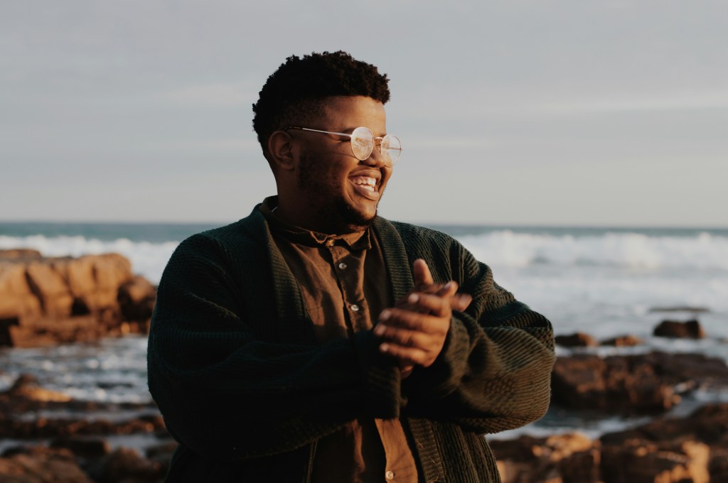A photo by Ethan Smith, featuring a Black man the photographer calls Selby, in a larger body with short hair and round, wire-framed glasses, smiling with his hands clasped together as he stands upon a rocky beach, with a small and distant ocean wave headed toward the shore. He is wearing a dark brown corduroy sweater and underneath it, a warmer brown button-up with marble colored buttons. He looks off to the side, appearing joyful.
