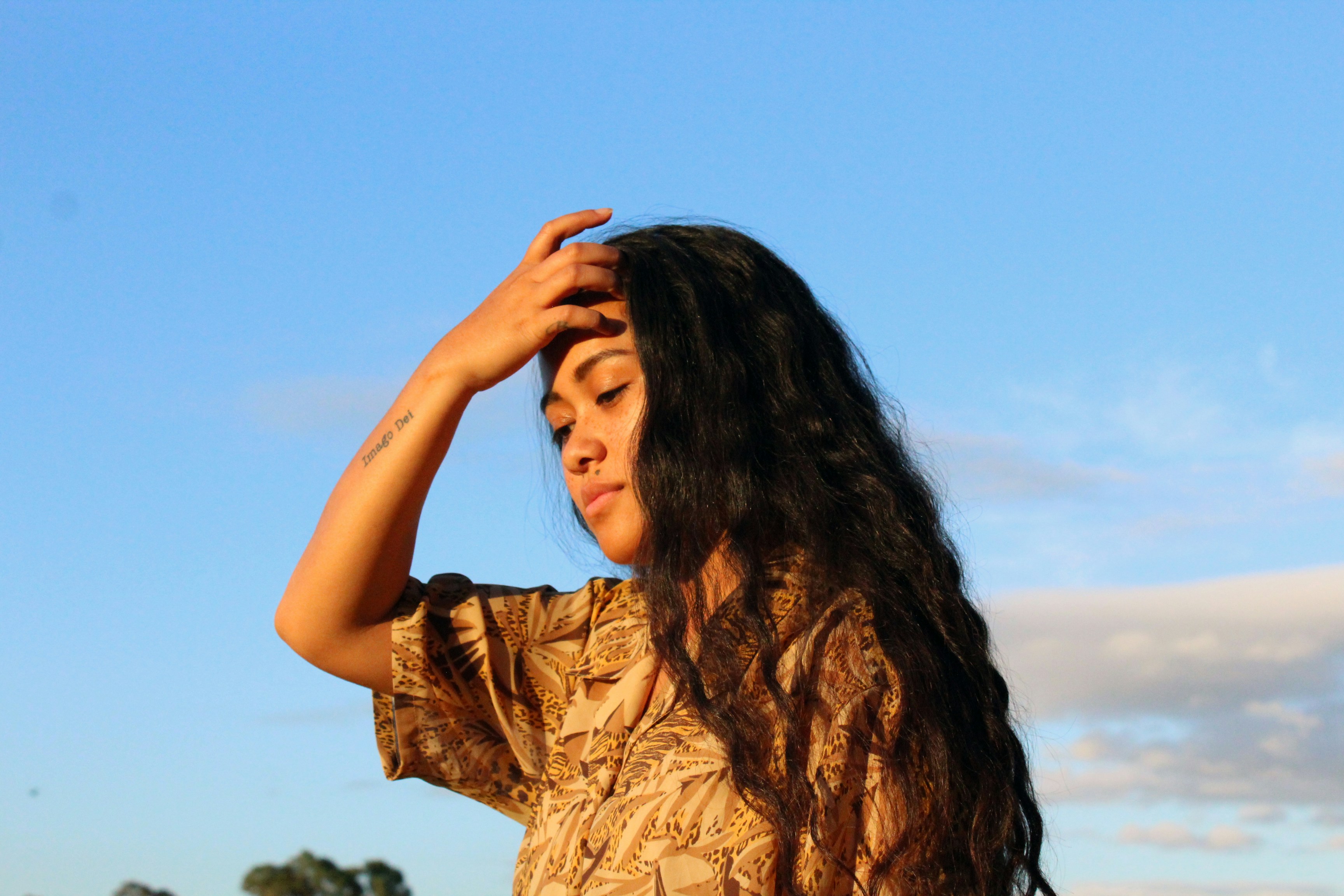 A photo by Gabrielle Henderson featuring a thin person with medium tan, glowing skin and dark, wavy hair, who stands against a bright blue sky, with their hand holding back their long hair. Their arm features a tattoo that says "imago dei," translating to "image of God," honoring the divine nature of human beings. They are wearing a short-sleeved mustard button-up with a slightly darker, paisley pattern. Their eyes are cast downward, as if peacefully contemplating something.