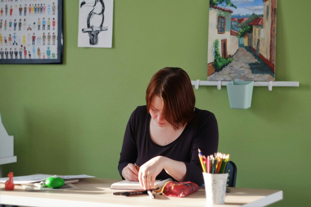 A light-skinned person sitting at an art table, creating something with a marker and a notepad.